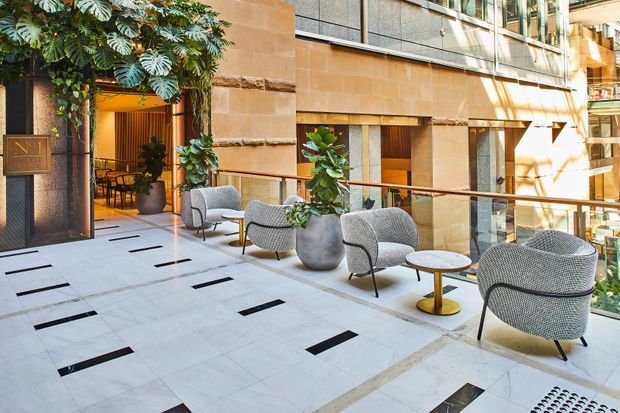 A refined open-air commercial atrium or terrace with pale stone paving, black inset floor accents, warm sandstone walls, glazed upper levels, and contemporary outdoor lounge seating arranged around large planters. A restaurant or hospitality space is visible through the entrance, with abundant greenery integrated into the setting.