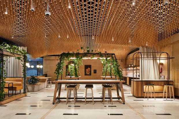 A spacious contemporary hospitality or commercial interior with a large communal timber table and stools, booth seating, shelving, and abundant hanging greenery. The room features a highly distinctive sculptural timber slatted ceiling with integrated lighting, pale stone or tile flooring with dark rectangular inlays, warm wood joinery, and a neutral beige palette. The open-plan layout has a refined, inviting atmosphere and appears designed for communal dining or work.