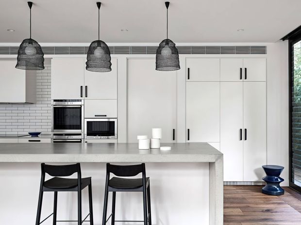 Bright contemporary kitchen with a large pale stone island, white full-height cabinetry, integrated stainless-steel ovens, white subway-tile splashback, and three black woven pendant lights. Black bar stools sit at the island, while dark timber flooring and a glazed opening add contrast.