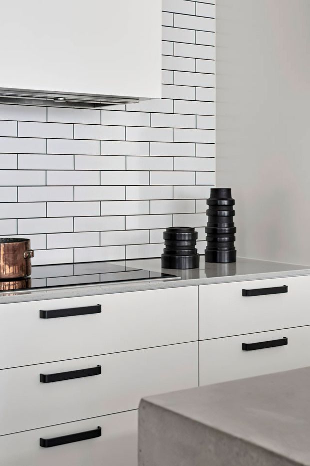 Close-up view of a contemporary kitchen with white lower cabinetry, black bar pulls, a black induction cooktop, white subway-tile splashback with dark grout, and a stainless-steel rangehood. Dark decorative vessels and a copper pot sit on the cooktop and benchtop, while a concrete-look island edge is visible in the foreground.