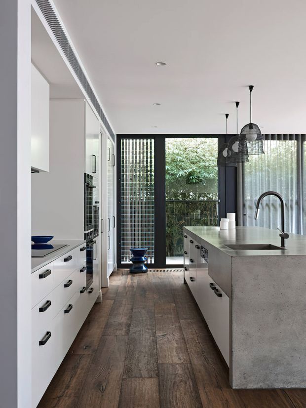 A contemporary galley-style kitchen with extensive white built-in cabinetry and black handles, a large central island with a grey concrete-look benchtop and integrated sink, dark timber-look flooring, and black-framed glazed doors and windows opening to greenery. Clustered black pendant lights hang above the island, while sheer curtains and vertical screening soften the rear glazing.