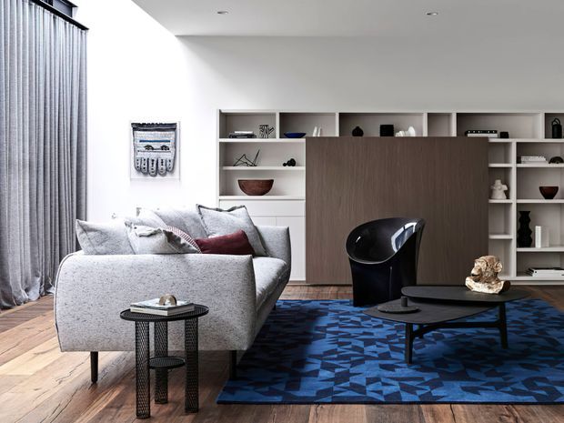Contemporary living room with a grey upholstered sofa, dark sculptural lounge chair, black oval coffee table, and a blue geometric area rug. Built-in white shelving and dark timber cabinetry form a feature storage wall, complemented by full-height grey curtains and timber flooring.