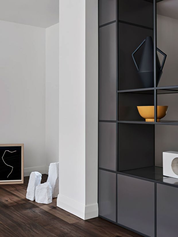 A minimalist interior corner featuring a tall dark built-in shelving and cabinetry unit with open display compartments. Sculptural black, white, and ochre objects are arranged on the shelves. A white structural column and wall meet a dark timber floor, with abstract artwork and white sculptural forms visible at the side.