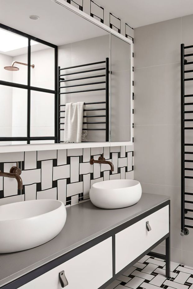 Contemporary bathroom featuring a double vanity with white vessel basins, a grey benchtop, white cabinetry with dark trim, bronze-toned wall-mounted taps, black heated towel rails, and bold geometric black-and-white tiled splashback and flooring. A large mirrored cabinet and black-framed shower glazing add strong graphic contrast.