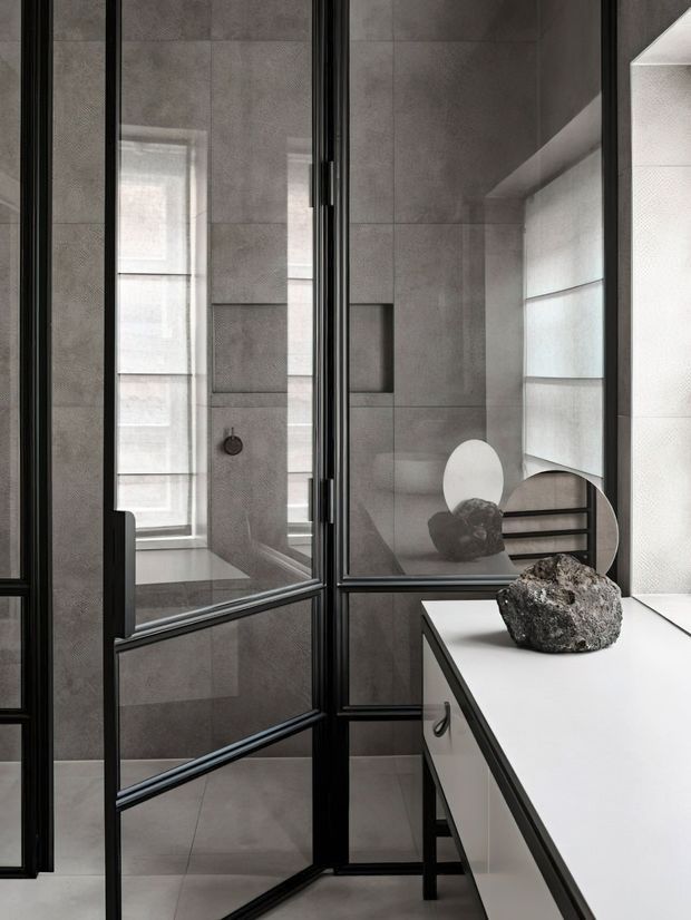 A refined grey-toned bathroom viewed through black-framed glass partition doors. Large-format grey wall and floor tiles, a wall-mounted shower fixture, frosted or translucent window treatments, and a white built-in vanity with dark edging are visible. A sculptural stone object sits on the vanity, while circular mirrors and reflections add depth. The space has a minimalist, contemporary appearance with strong geometric lines and natural muted materials.