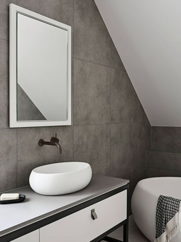 A compact contemporary bathroom beneath a sloped ceiling, featuring large grey textured wall tiles, a white vessel basin on a dark-edged white vanity, a wall-mounted dark metal tap, a framed mirror, and a partially visible freestanding white bathtub. The palette is neutral grey, white, and black with clean modern lines.