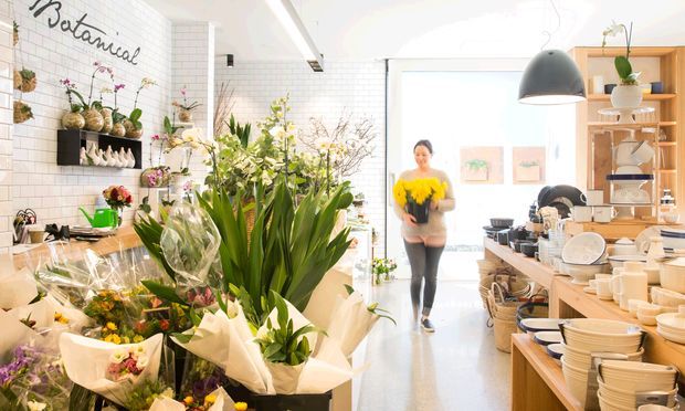 Bright botanical-themed retail interior combining a flower shop and homewares display. White tiled walls, abundant potted plants and cut flowers, open timber shelving, ceramic tableware, and a large black pendant light define the space. A wide glazed entrance at the rear brings in strong daylight.