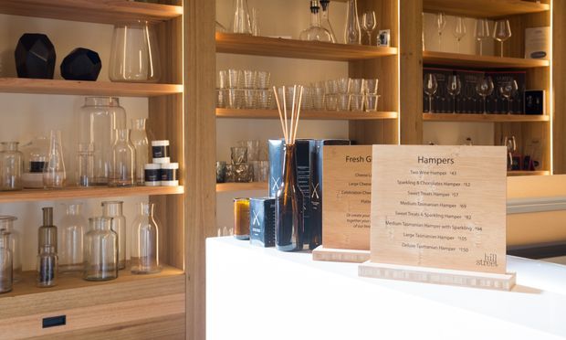 A warmly lit retail or hospitality display area with open timber shelving filled with glassware, decanters, bottles, and boxed products. A white counter in the foreground presents fragrance or gift products alongside printed menus or promotional signs. The scene has a refined, curated showroom feel with integrated shelf lighting and extensive glass displayware.