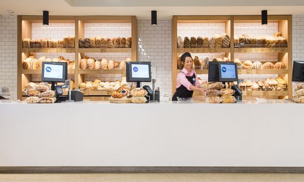 A bright bakery or bread-shop interior viewed from the customer side of a long white service counter. Behind the counter are multiple timber-framed shelving bays with warm lighting, white subway-tile wall backing, and shelves stocked with assorted breads and pastries. Several point-of-sale monitors and a staff member are visible. The space has a clean, contemporary commercial fit-out with neutral finishes and warm wood accents.