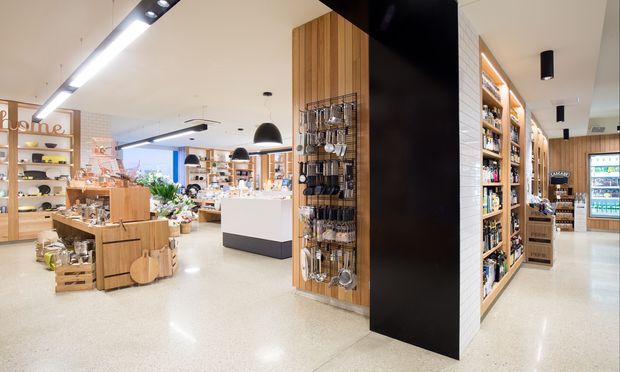 Bright contemporary retail interior with polished light terrazzo flooring, white ceilings, timber display shelving and cabinetry, black feature joinery, and integrated linear and pendant lighting. The open-plan shop floor contains product displays, shelving, checkout or service counters, and a refrigerated display area at the rear.