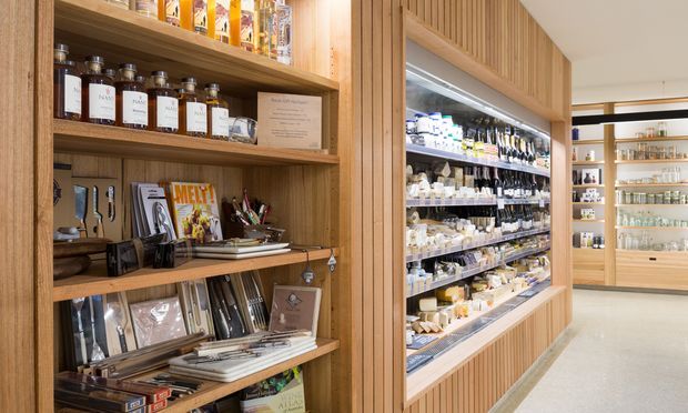 A warmly lit retail interior featuring timber shelving and extensive built-in display cabinetry. Shelves hold packaged goods, jars, bottles, books, magazines, and gift items, while a long illuminated refrigerated or glass-front display case occupies the centre-right. Vertical timber battens and pale wood finishes create a refined, organised shop fit-out.