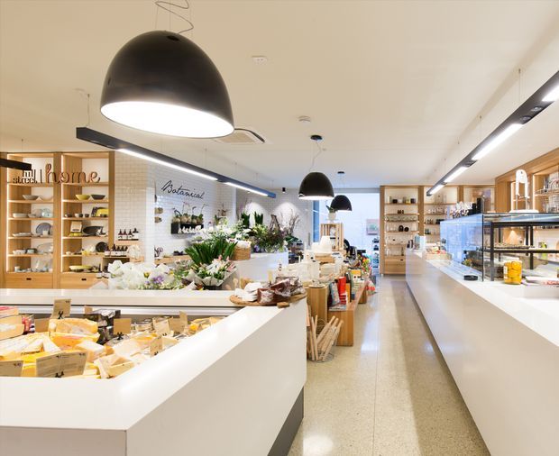 Bright contemporary specialty food shop interior with long white service counters, glass display cases, timber open shelving, exposed white brick, abundant floral and food displays, and large black pendant and linear ceiling lights. The image is dominated by the retail/commercial fit-out and showcases a clean, refined food-market atmosphere.