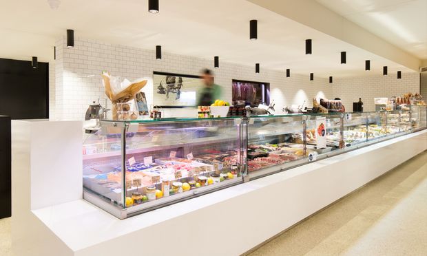 Bright contemporary commercial food shop or deli interior with a long refrigerated glass display counter filled with prepared foods and produce. The space has white-painted subway-tile walls, white built-in counter cabinetry, pale flooring, and numerous black cylindrical pendant lights arranged across the ceiling. Staff and food displays are visible behind the counter.