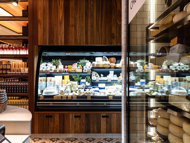A richly detailed specialty food retail interior featuring a large refrigerated display case built into dark timber wall panelling. Shelves display cheeses, packaged foods, bottles, and other gourmet products, with additional illuminated shelving and reflected merchandise visible on the right. Warm timber, white tiled walls, black metal framing, integrated lighting, and patterned floor tiles create a refined, warm commercial fit-out.