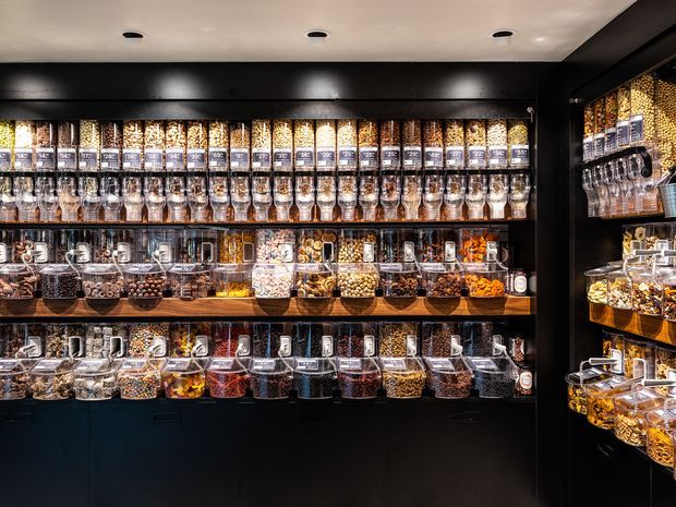 A compact retail bulk-food display wall with multiple rows of clear dispensers, jars, and containers filled with cereals, grains, nuts, sweets, and dried foods. The shelving and cabinetry are predominantly black, with warm integrated lighting highlighting the products. The image focuses on the built-in merchandising joinery and organized commercial interior.