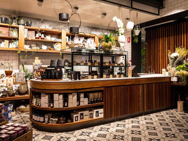 A warmly lit retail café or specialty shop interior centred on a curved timber service counter with open shelving and a glass display cabinet. The counter and shelving display packaged goods, bottles, crockery and decorative items. Vertical dark timber slats frame the right side, while white subway-tile walls, black metal detailing, hanging pendant lights, and patterned floor tiles create a refined, boutique atmosphere.