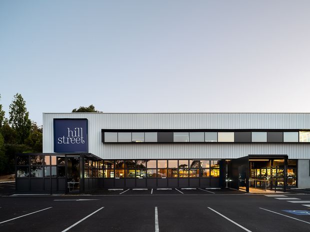 Exterior view of a contemporary two-storey commercial building at dusk, with a long corrugated metal facade, horizontal ribbon windows, dark-framed glazed ground-floor frontage, a projecting entrance canopy, and a prominent 'hill street' sign. The building fronts a marked asphalt car park and is bordered by trees.