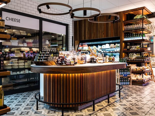A richly detailed specialty food shop or deli interior, centred on a curved dark wood retail counter with warm vertical slat detailing and integrated amber lighting. Behind it are refrigerated cheese and delicatessen displays, while open timber shelving holds bottles, jars and packaged goods. The space combines white subway-tile walls, dark-framed glazing, dark timber wall panelling, patterned geometric floor tiles and circular black pendant lighting. The image is bright, warm and highly layered, with a polished hospitality-retail character.