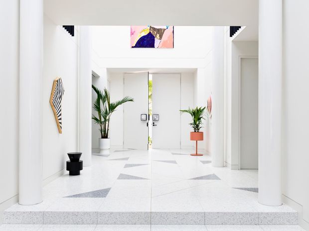 Bright, minimalist entrance foyer with white walls, large cylindrical columns, terrazzo flooring and geometric grey inlays. Double doors open to a narrow planted lightwell, while potted palms, sculptural wall art and a small black pedestal add restrained decorative accents. The space has a high ceiling and strong natural light.
