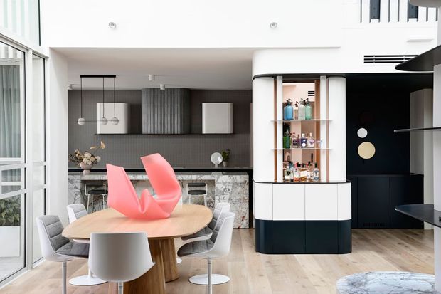 Bright open-plan contemporary kitchen and dining area with a sculptural timber dining table, modern white chairs, a dark tiled kitchen backsplash, stone-patterned island cabinetry, and a striking curved built-in drinks cabinet. Floor-to-ceiling glazing and pale timber flooring create an airy, refined interior.