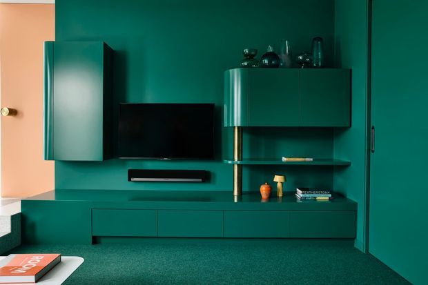 Bold, monochromatic teal-green media wall with streamlined built-in cabinetry, a wall-mounted television, soundbar, floating shelves, and a long low console. A tall cupboard and doorway frame the composition, while a peach-toned adjacent wall and small decorative objects provide contrast. The room has a richly saturated, refined contemporary feel.