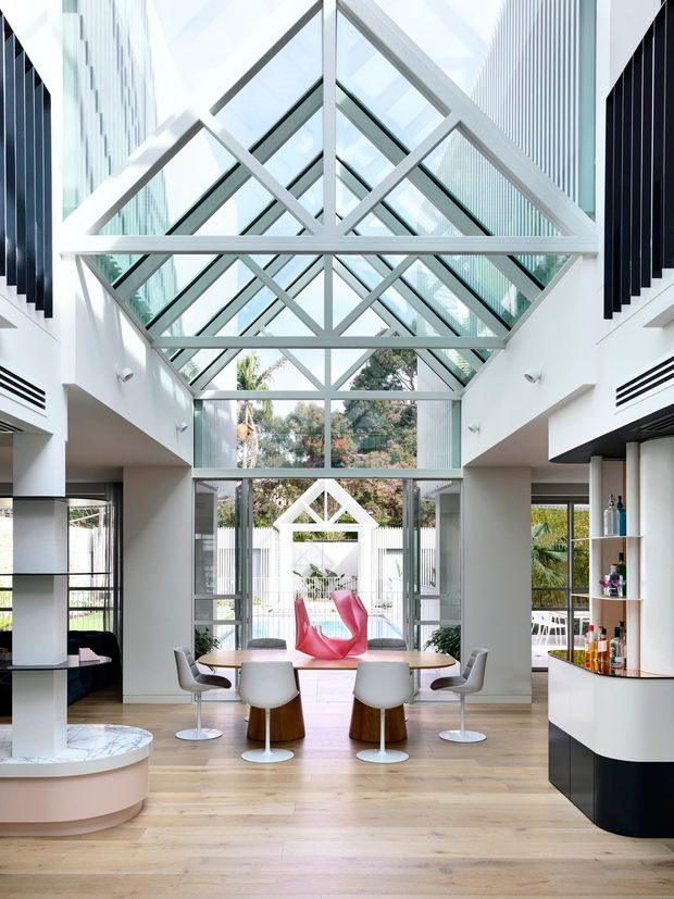A bright, contemporary open-plan interior centred on a dramatic double-height glazed atrium. A large white-painted structural frame forms repeated triangular and diamond-shaped geometric patterns beneath a glazed pitched roof, bringing abundant daylight into the space. The room combines a dining area with a sculptural table and modern pedestal chairs, a curved built-in bar or kitchen area with open shelving, and a rounded marble-topped feature bench in the foreground. Floor-to-ceiling glazing opens toward an outdoor courtyard or pool area, while bold pink lounge seating provides a vivid accent. The palette is predominantly white, pale timber, light grey, and glass, with strong geometric architectural detailing.