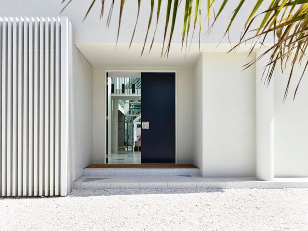 A minimalist residential entrance with a deep navy pivot door set within a white recessed entry. Vertical white screening forms the left side of the facade, while a broad white column frames the right. Pale stone steps and a light gravel forecourt lead to the doorway, with tropical foliage overhead. The open door reveals a bright interior beyond.