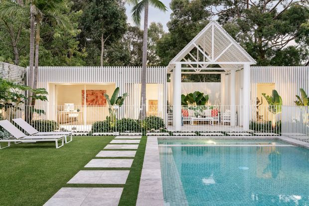 A bright tropical courtyard with a rectangular swimming pool, pale stone paving, manicured lawn, and a stepping-stone path. A white vertical-slat boundary fence encloses the space, with a distinctive open-sided white pavilion featuring a steep gabled roof and decorative latticework. Dense palms and greenery surround the pool area, with white loungers and planted garden beds adding to the relaxed resort-like setting.