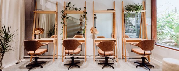 A bright, warm-toned salon or beauty studio with four individual styling stations. Each station has a timber-framed mirror, floating shelf, and brown upholstered swivel chair, with trailing plants and a large window adding a natural, relaxed atmosphere. Light flooring, neutral walls, and timber detailing create a contemporary organic interior.