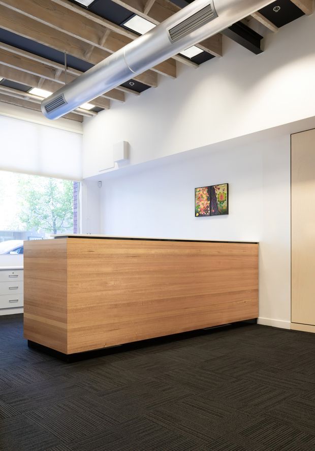 Bright modern commercial reception area with a large fixed timber reception desk, dark modular carpet flooring, white walls, exposed timber ceiling beams, black ceiling panels, recessed lighting, and a prominent silver cylindrical HVAC duct. A large window provides natural light, with a small artwork and pale built-in cabinetry visible.
