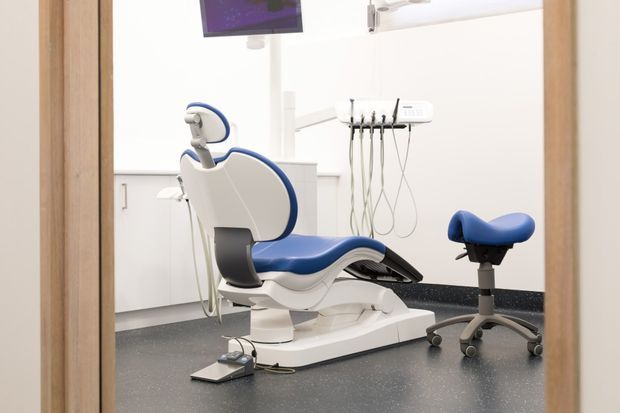A clean, modern dental treatment room viewed through a timber-framed doorway. The room contains a white dental chair with blue upholstery, overhead dental lighting, wall-mounted dental instruments, a matching adjustable dentist's stool, white cabinetry, and dark speckled flooring with a dark backsplash.