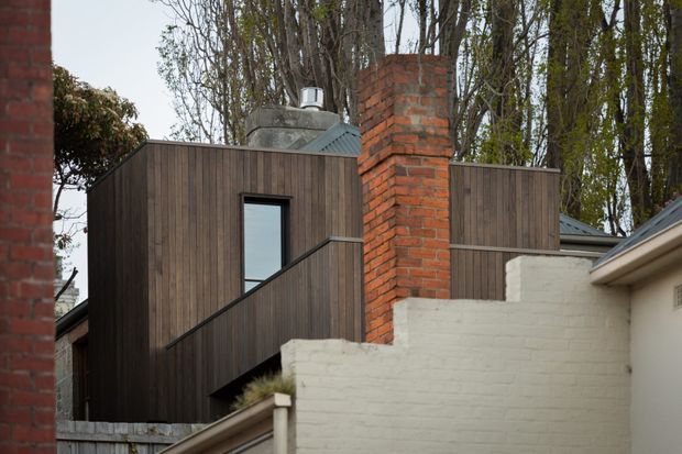 Exterior architectural view of a contemporary extension or dwelling, featuring dark vertical timber cladding, a small black-framed window, a sloping metal roof, and a prominent red brick chimney. A white-painted brick wall and neighbouring rooflines occupy the foreground, with mature trees behind the building.