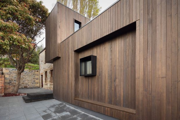 Exterior view of a contemporary timber-clad building or extension in a landscaped courtyard. The facade features vertical dark natural-wood boards, sharp projecting and recessed forms, black-framed windows, and a small projecting canopy or overhang. Paved hardscaping leads past an older rough stone wall and mature planting, creating a contrast between the modern addition and the existing masonry structure.