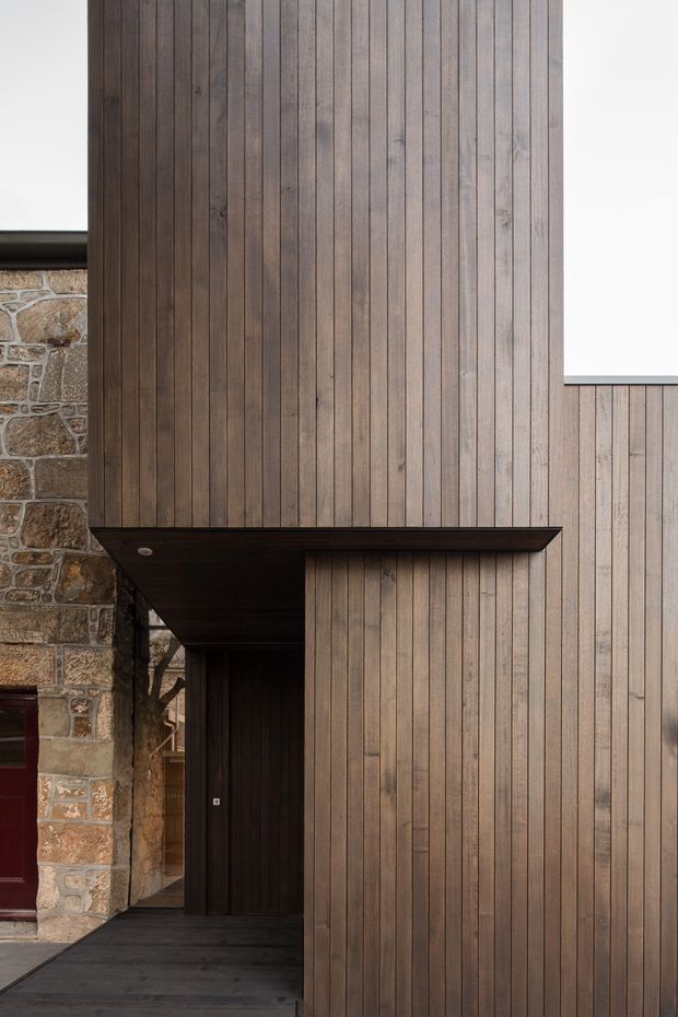 A close architectural view of a contemporary entrance facade, featuring vertically aligned dark timber cladding, a deep projecting canopy, a recessed dark timber entry door, and contrasting irregular natural stone masonry at the side. The composition emphasizes material texture, strong vertical lines, and clean modern detailing.