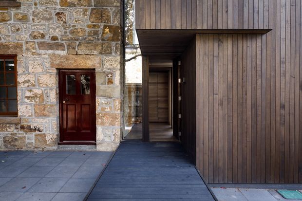 Exterior architectural view showing a contemporary timber-clad addition connected to an older stone building. The composition contrasts dark vertical timber boarding and a recessed covered entry with the original rough stone masonry and dark wood door. Grey paved surfaces lead toward the entrance, creating a strong juxtaposition between historic and modern construction.