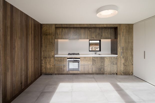 Minimal, contemporary kitchen with full-height dark timber-look wall panelling and cabinetry, a compact island-free cooking zone, integrated stainless-steel oven and cooktop, pale smooth splashback, and large light-grey tiled flooring. Tall white storage doors occupy the right side, while a flush circular ceiling light and clean-lined detailing reinforce the restrained modern design.