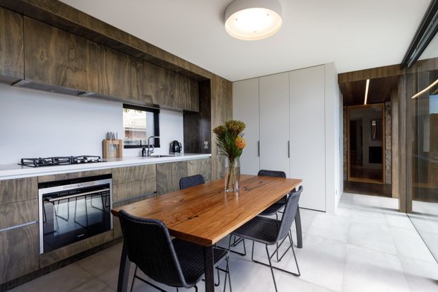 Contemporary open-plan kitchen and dining area with dark timber cabinetry and wall panelling, white benchtops, integrated cooking appliances, a large natural timber dining table, and black woven dining chairs. Full-height glazing and a timber-lined hallway add depth, while white cabinetry and pale flooring keep the space bright.