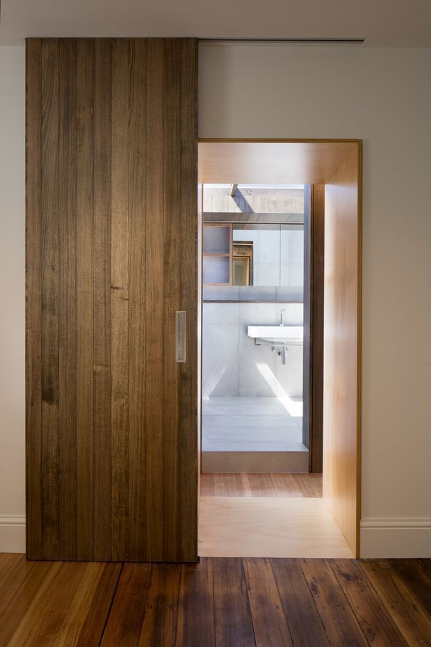 Interior view of a warm, dark timber-lined sliding door or concealed wall panel opening onto a compact contemporary bathroom. The bathroom features pale stone or tiled surfaces, a wall-mounted white basin, mirrored shelving, and a timber-framed doorway. Dark timber flooring continues through the foreground, contrasting with the light bathroom interior and neutral painted walls.
