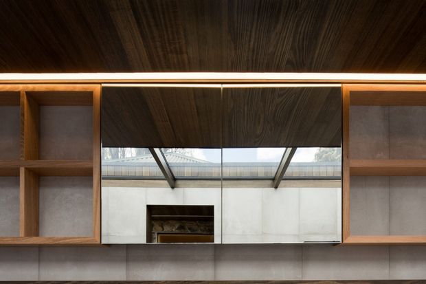 Close-up architectural interior detail of a dark timber-lined built-in wall and ceiling, with symmetrical open timber shelving, pale integrated cabinetry or wall panels, concealed linear lighting, and a central mirrored or glazed section reflecting a skylit courtyard-like space and fireplace opening. The composition is refined and highly geometric, with warm timber contrasting against cool light-grey surfaces.