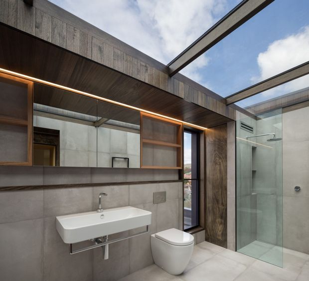 Contemporary bathroom with large-format grey concrete-look wall and floor tiles, a wall-mounted white basin and toilet, and a frameless glass shower enclosure. Warm timber cabinetry and ceiling lining contrast with dark metal detailing. A large mirror, integrated linear lighting, and overhead glazing create a refined, bright, modern space.