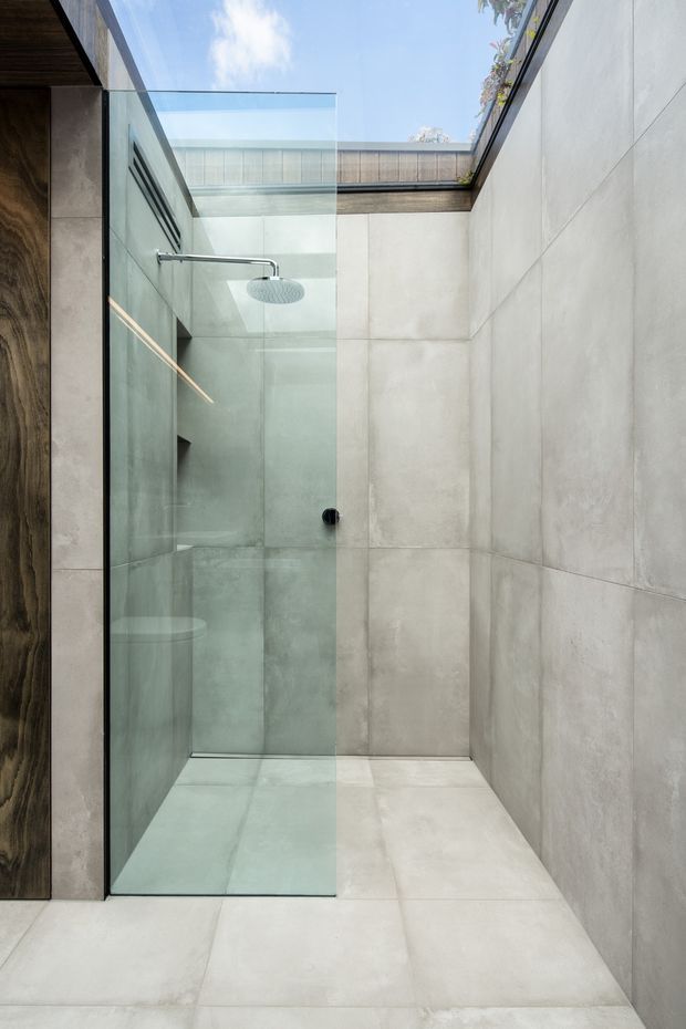 Minimalist walk-in shower in a contemporary bathroom, featuring large pale grey concrete-look wall and floor tiles, a frameless clear glass shower screen, a chrome rain shower, and a timber-toned vertical wall panel at left. An overhead skylight or open roof section brings in natural light and greenery is faintly visible above.