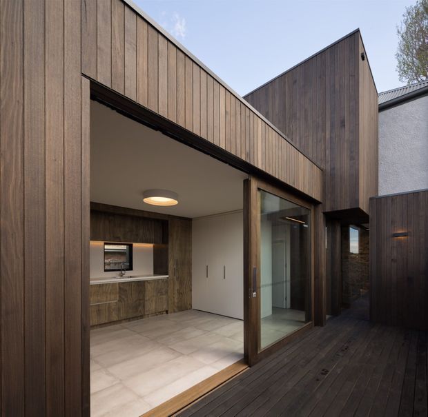 Exterior view of a contemporary courtyard-like residential addition clad in dark vertical timber boards. A covered open entry or outdoor living area leads into a compact interior with timber cabinetry, pale tiled flooring, a white flush door, and large glazed sliding doors opening onto a dark timber deck. The building has angular pitched roof forms and a restrained material palette.