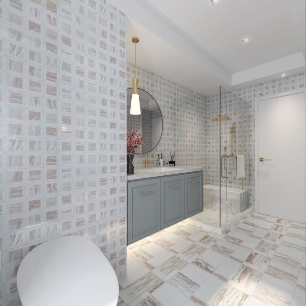 Bright contemporary bathroom with a frameless glass shower enclosure, pale grey-blue vanity with brass pulls, white stone benchtop, round wall mirror, and distinctive patterned marble-look wall and floor tiles in soft white, grey, and blush tones. A freestanding bathtub is partially visible in the foreground.