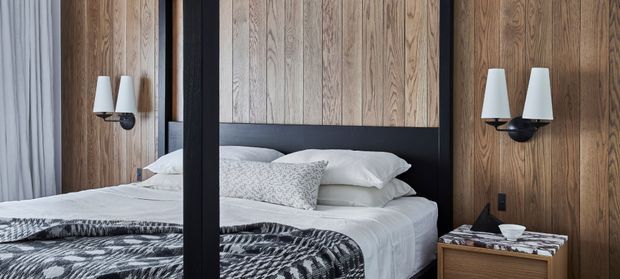 A contemporary bedroom with a bed framed by dark timber or metal posts, vertical warm wood wall panelling, black wall-mounted sconces, and layered white and grey bedding. A full-height mirror reflects the bed and curtains, while a timber bedside table adds warmth.