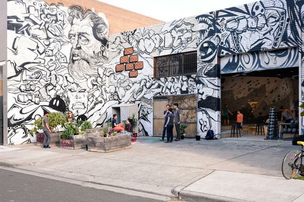 Exterior view of a large urban commercial or industrial-style building with an expansive black-and-white graffiti mural covering the facade. The building has exposed red brick sections, a dark metal-framed window, a wide open entrance, and a terrazzo-like geometric wall surface visible inside. Planter boxes and several people occupy the paved forecourt.