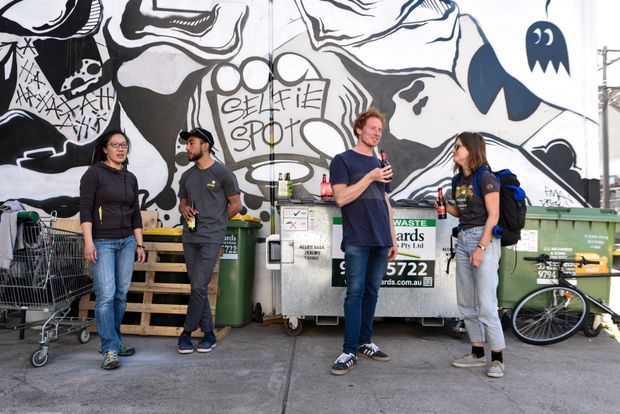 An outdoor urban service area beside large waste and recycling bins, with four people standing in front of a bold black, white, and blue abstract mural. Shopping carts, a bicycle, pallets, and utility infrastructure are visible around the concrete-paved space. The image is documentary in style and primarily depicts a commercial or public-facing waste collection area rather than an interior architectural space.