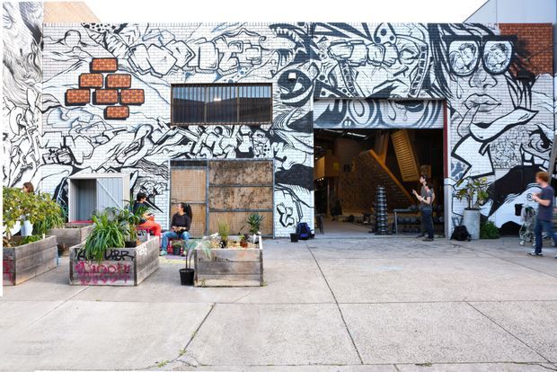 A wide exterior view of an urban commercial or community building with a large black-and-white illustrated mural covering the facade. The concrete forecourt contains raised timber planter boxes with greenery and informal seating, while a tall open entrance reveals an industrial-looking interior with stairs and exposed brick. People are present around the entrance, and portions of corrugated or metal cladding and brick are visible behind the artwork.