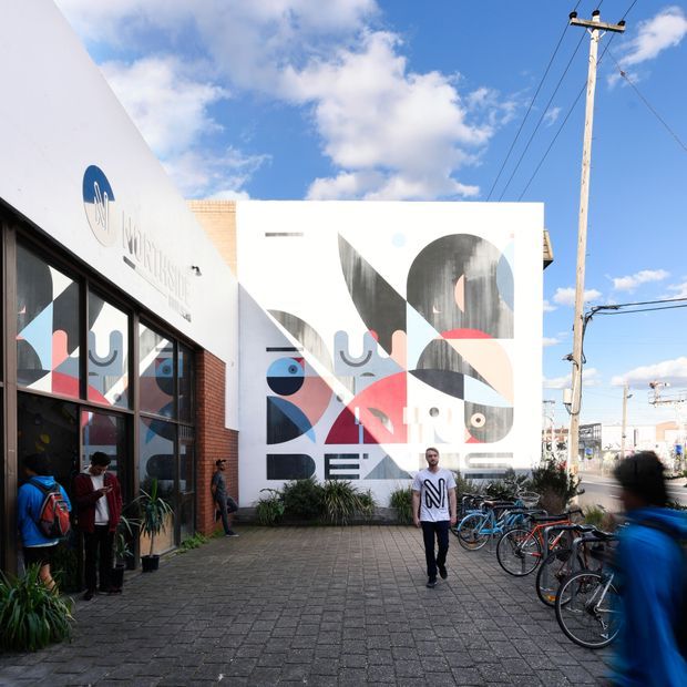 A bright urban exterior courtyard beside a contemporary low-rise building, featuring a large geometric abstract mural in black, white, blue, pink, and grey on a white facade. The foreground is paved with dark concrete blocks and includes bicycles, pedestrians, potted plants, brick detailing, large glazed windows, and a timber utility pole. The scene has a colourful public-art and creative-campus character under a partly cloudy sky.