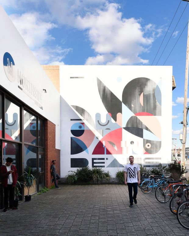Street-level view of a contemporary commercial building frontage and paved courtyard. A large geometric mural covers the white facade, with adjacent glazed shopfront windows, exposed brickwork, signage, bicycles, planting, pedestrians, and overhead utility lines under a bright partly cloudy sky.