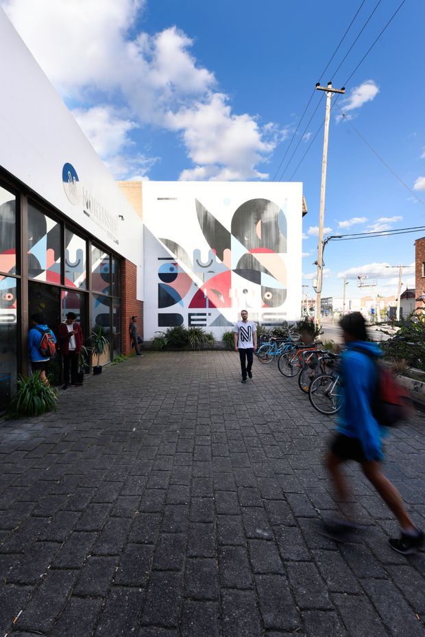 An urban pedestrian forecourt beside a contemporary low-rise commercial building, featuring a large geometric mural on a white facade, glazed shopfront windows, dark paver flooring, bicycles, and people using the space. Utility poles and neighbouring buildings are visible beyond the courtyard.